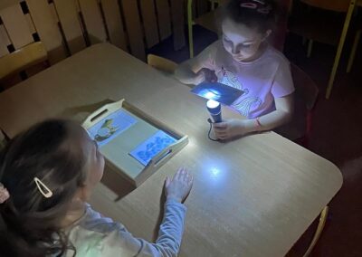 Dziewczynki podświetlają latarkami karty pracy.
